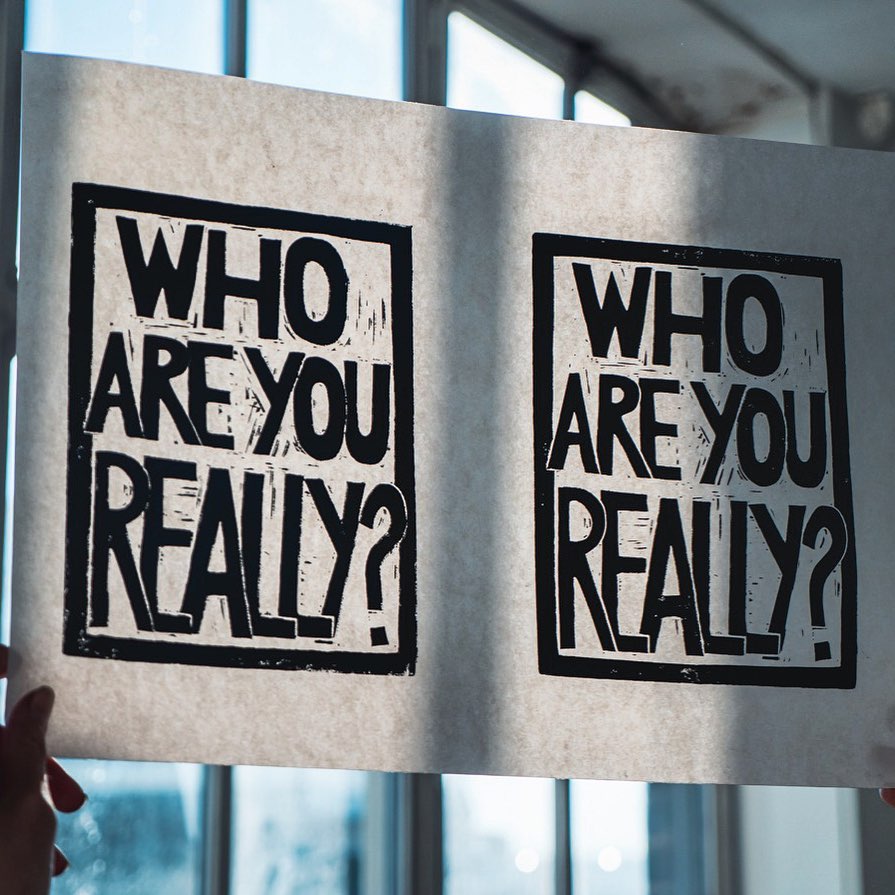 White printed poster with black border and block writing hangs in a window reads WHO ARE YOU REALLY?